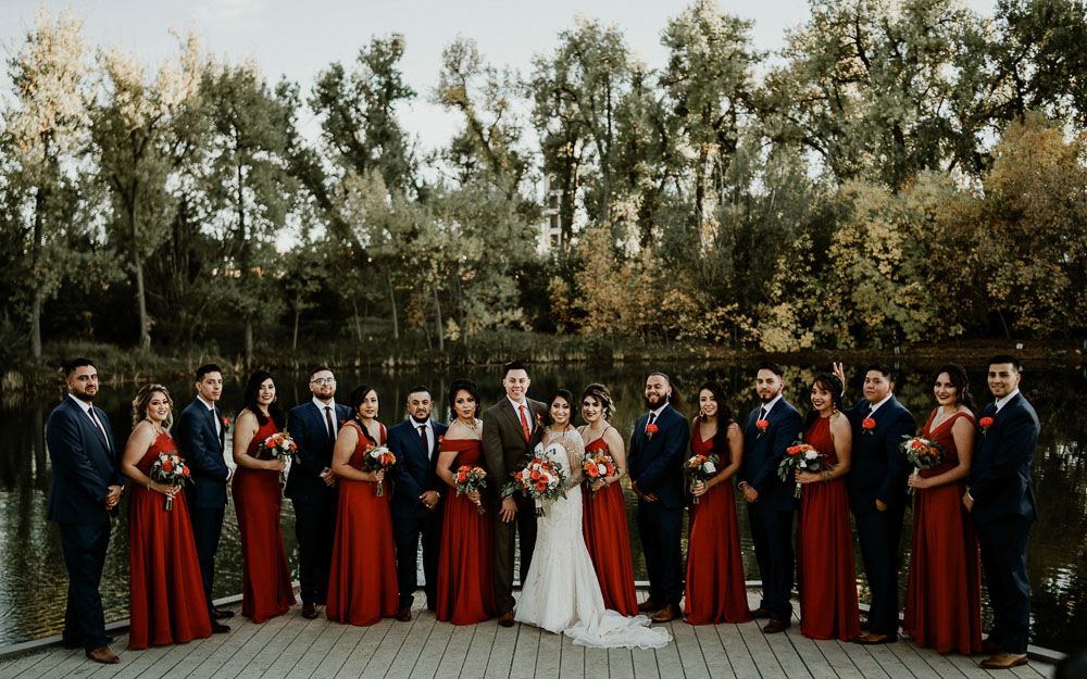 Large wedding party in red dresses and dark suits poses together outdoors near trees.