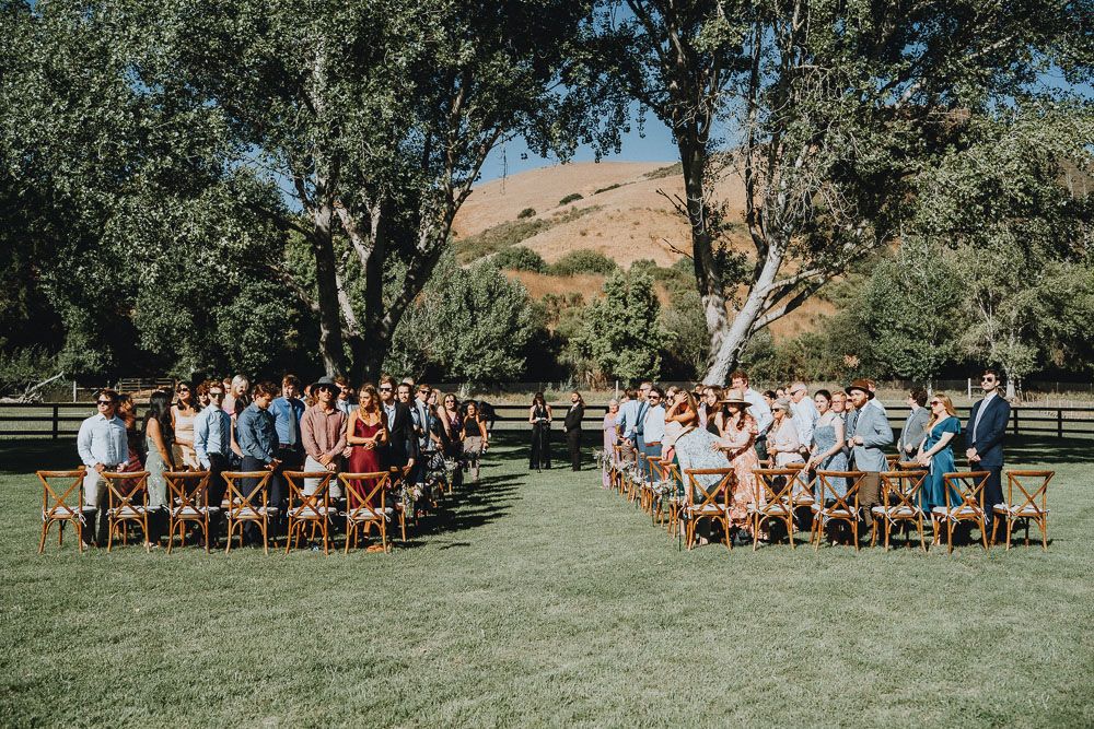 Outdoor wedding ceremony setup with wooden chairs under large trees.