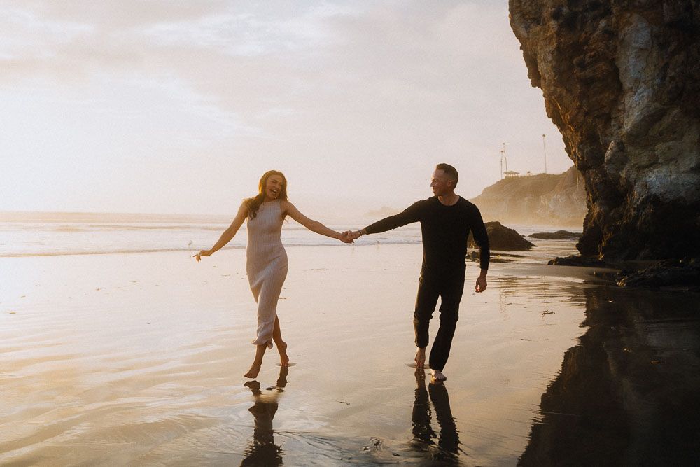 Silhouettes hold hands while walking along a misty beach at golden hour.