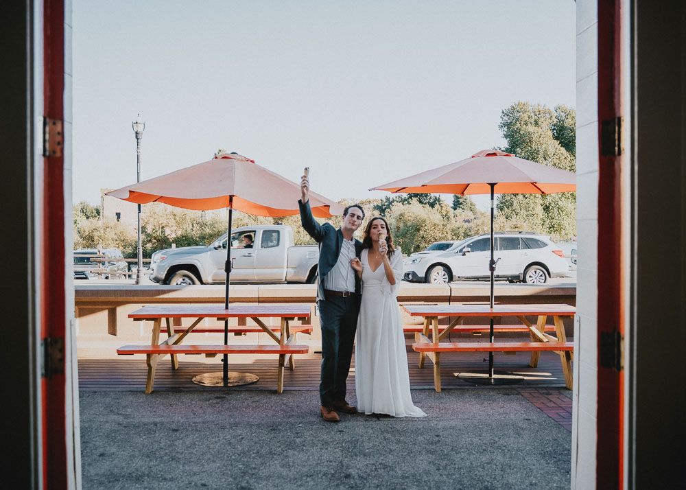 Newlyweds pose joyfully by orange patio umbrellas and picnic tables.