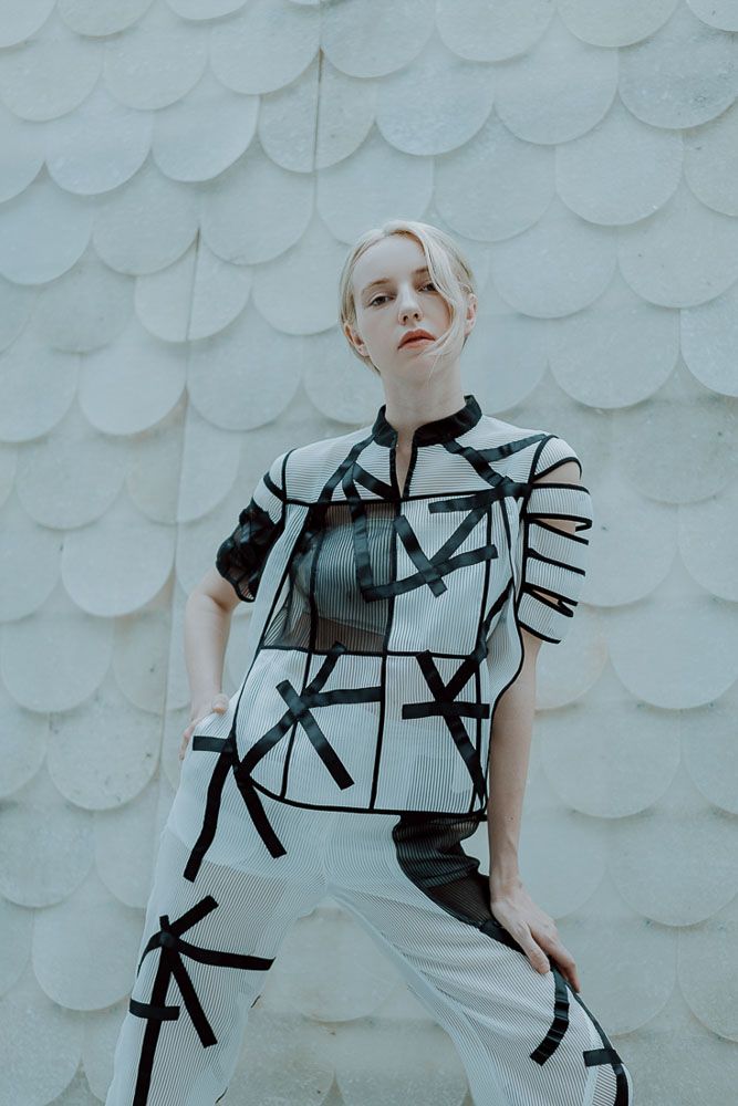 Model wearing geometric black and white patterned outfit against textured white wall.