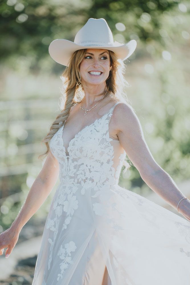 A bride wearing a white lace gown and cowboy hat poses outdoors in natural lighting.