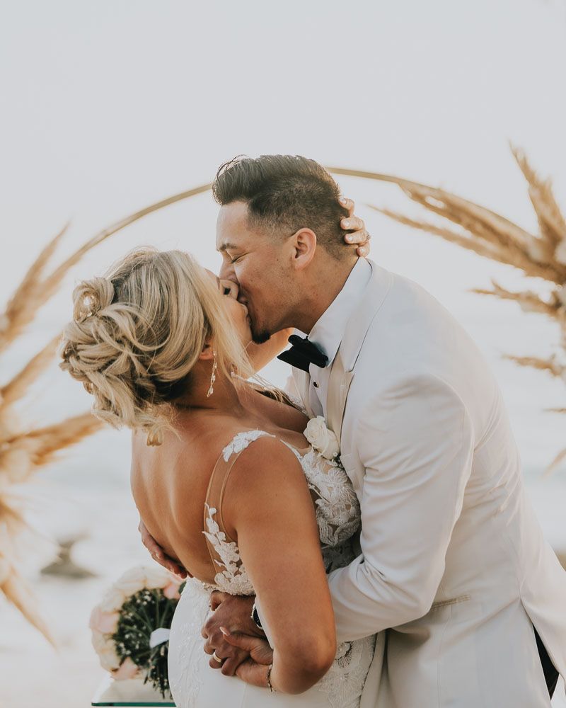 A tender moment between newlyweds against a soft dreamy beach background with pampas grass decor.