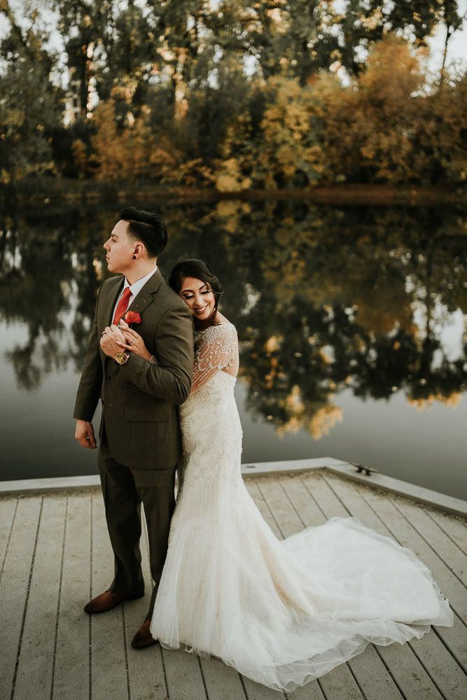Couple in wedding attire pose romantically on wooden dock by peaceful lake at sunset.