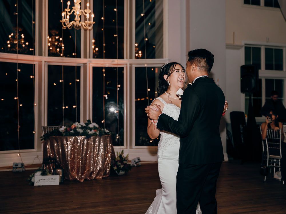 Couple shares their first dance at wedding reception with twinkling lights and windows in background.