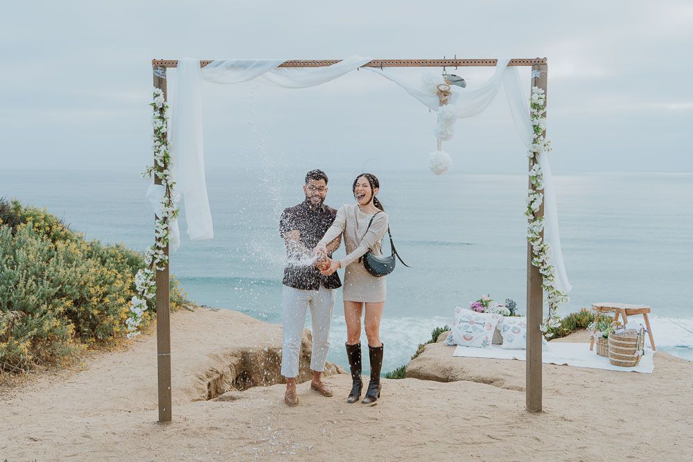 Couple stands under romantic beach arch decorated with white fabric and greenery overlooking the ocean.
