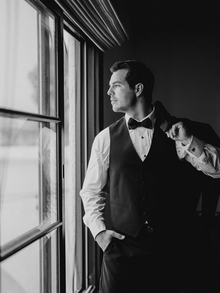 Groom in formal attire gazes thoughtfully out of window in dramatic black and white portrait.