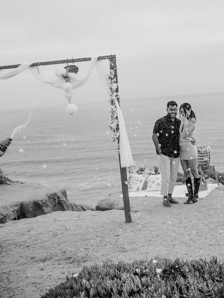 Couple stands by floral-decorated arch on coastal clifftop with ocean view.