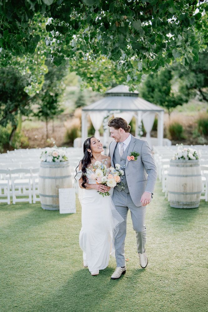 A romantic outdoor wedding scene with a couple on green lawn near wine barrels and white gazebo in background.