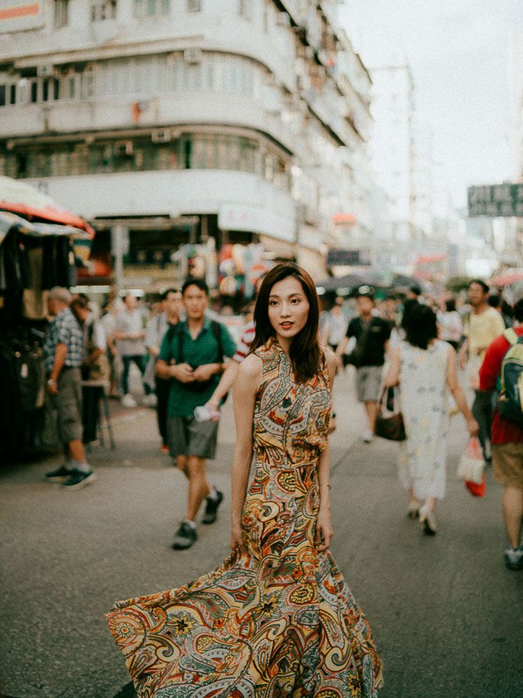 A woman in a flowing paisley dress stands in a bustling Asian street market scene.