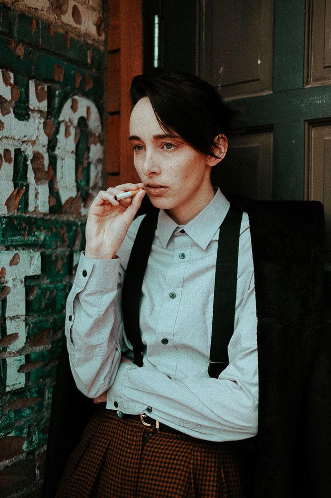 Figure in vintage-inspired outfit with white shirt, suspenders and plaid pants smoking in moody lighting.