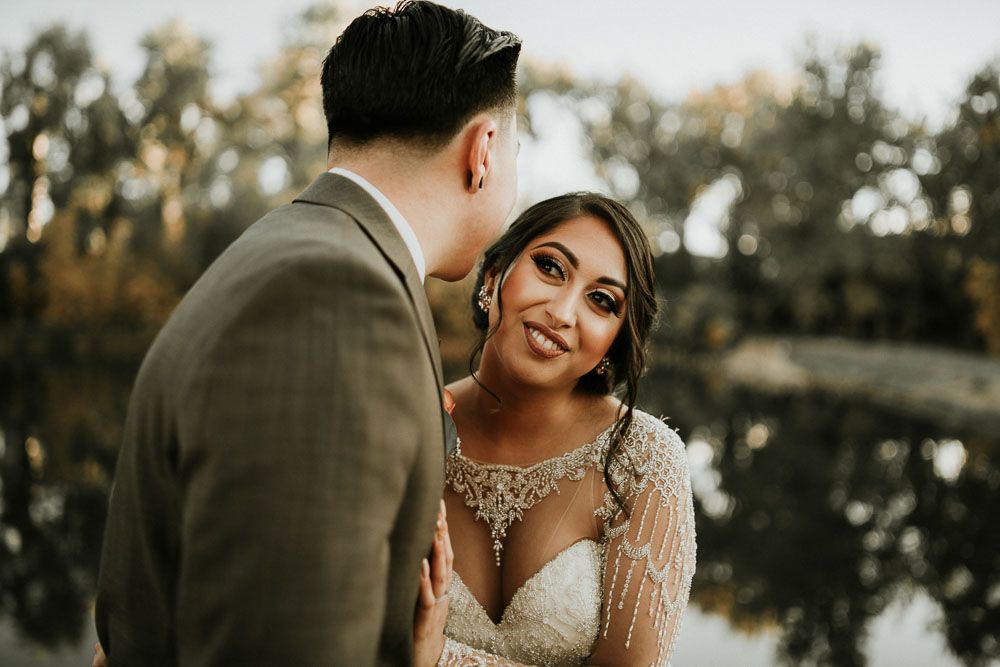 Intimate moment between couple in wedding attire with lake and trees in background.
