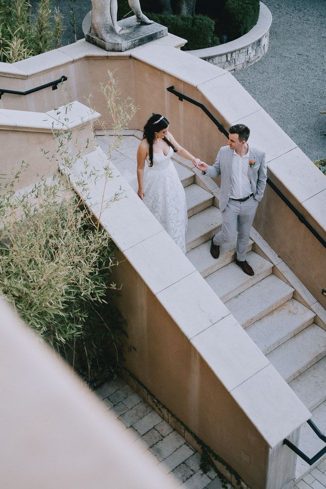 Wedding couple holding hands on curved outdoor staircase with Mediterranean architecture.