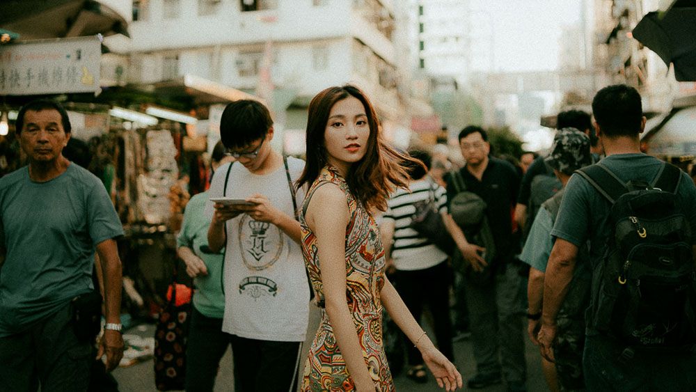 A woman in a floral dress stands amid a crowded Asian street market.