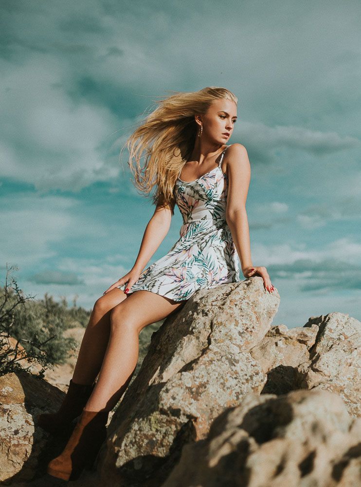Fashion model in floral dress poses on rocky outcrop against dramatic sky.