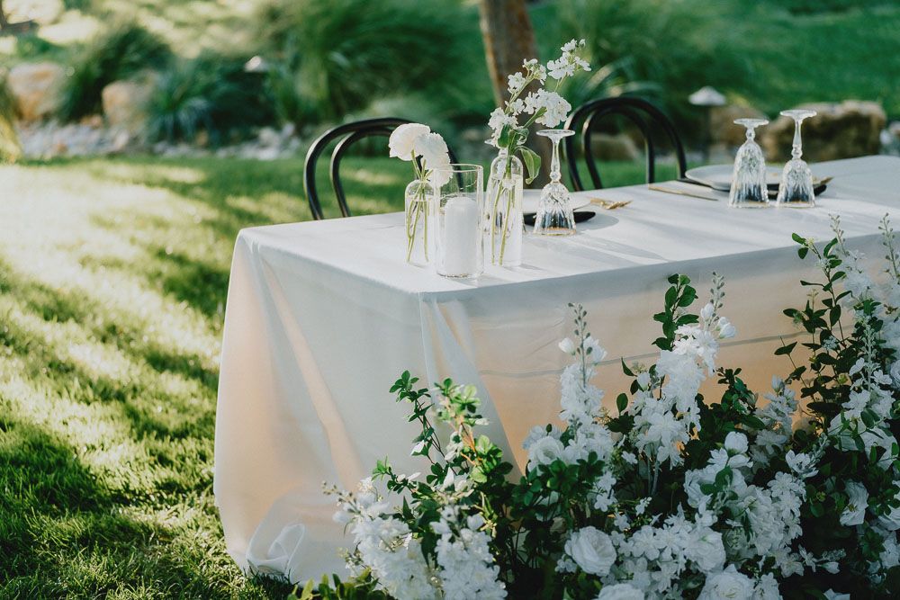 Wedding table setup with white linens and small vases of white flowers in natural outdoor lighting.