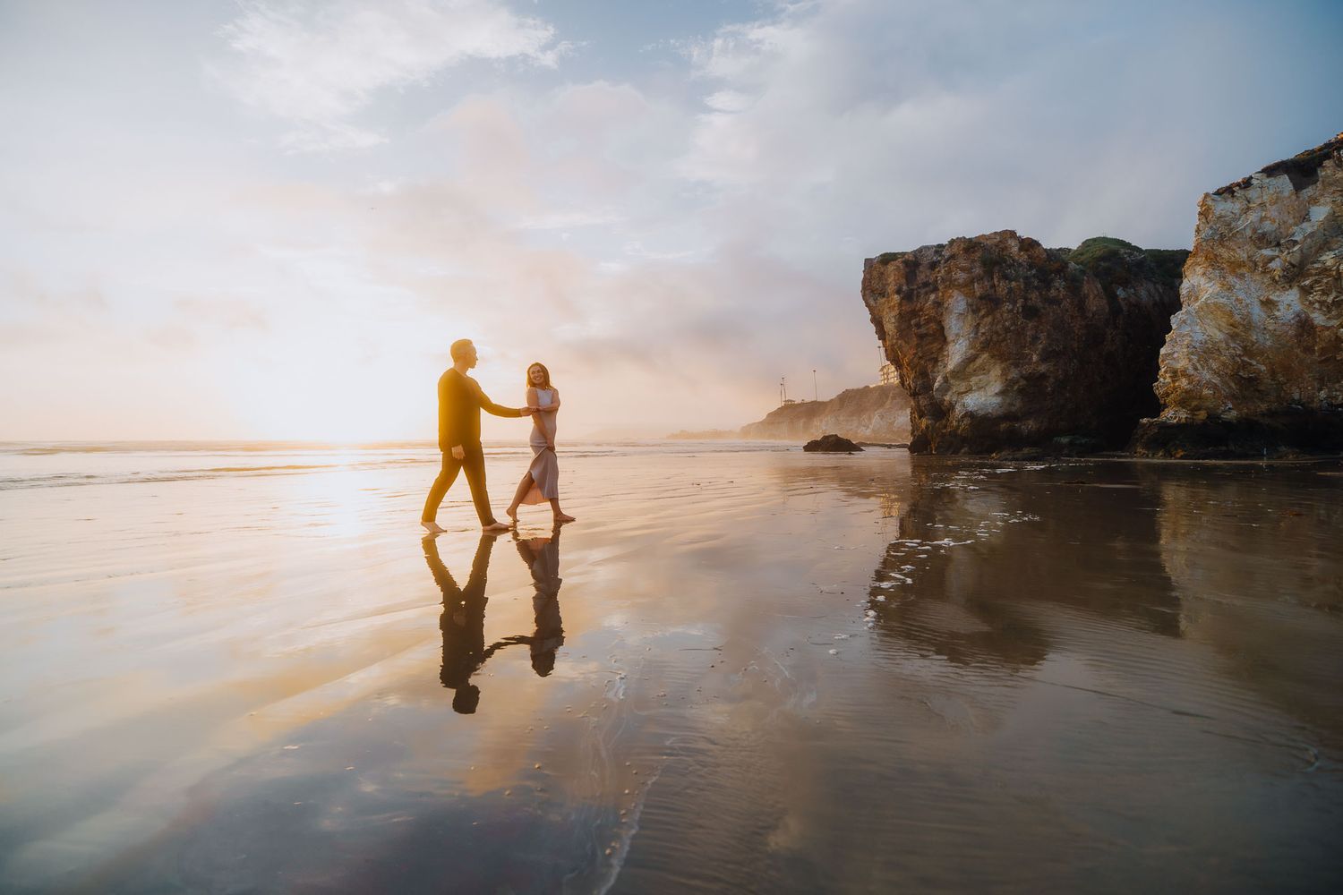 Silhouetted couple holding hands on reflective beach at sunset with coastal rocks in background.