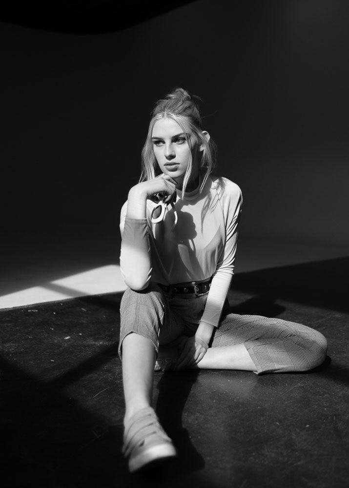 Fashion model in white top sitting on floor in black and white studio photograph.