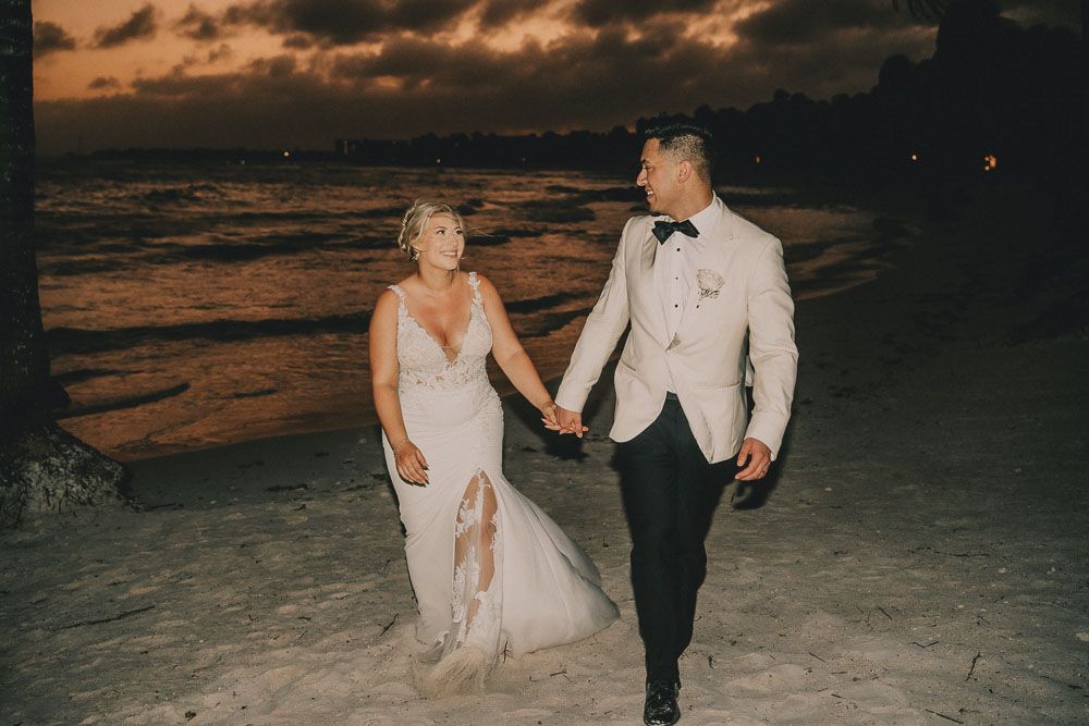 A wedding couple walks along a beach at sunset creating romantic silhouettes against the orange sky.