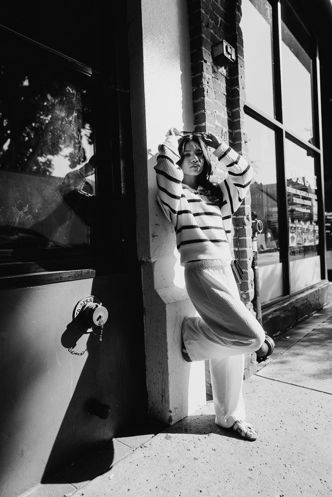 Black and white portrait against storefront featuring striped sweater and casual pose.