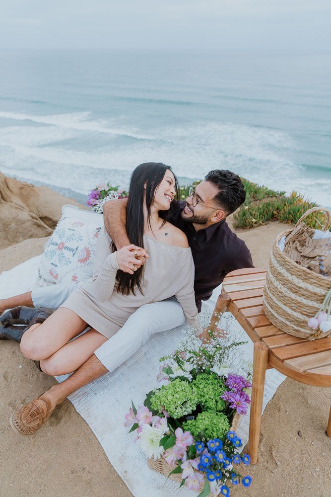 Romantic coastal picnic setup with flowers and couple enjoying ocean views.