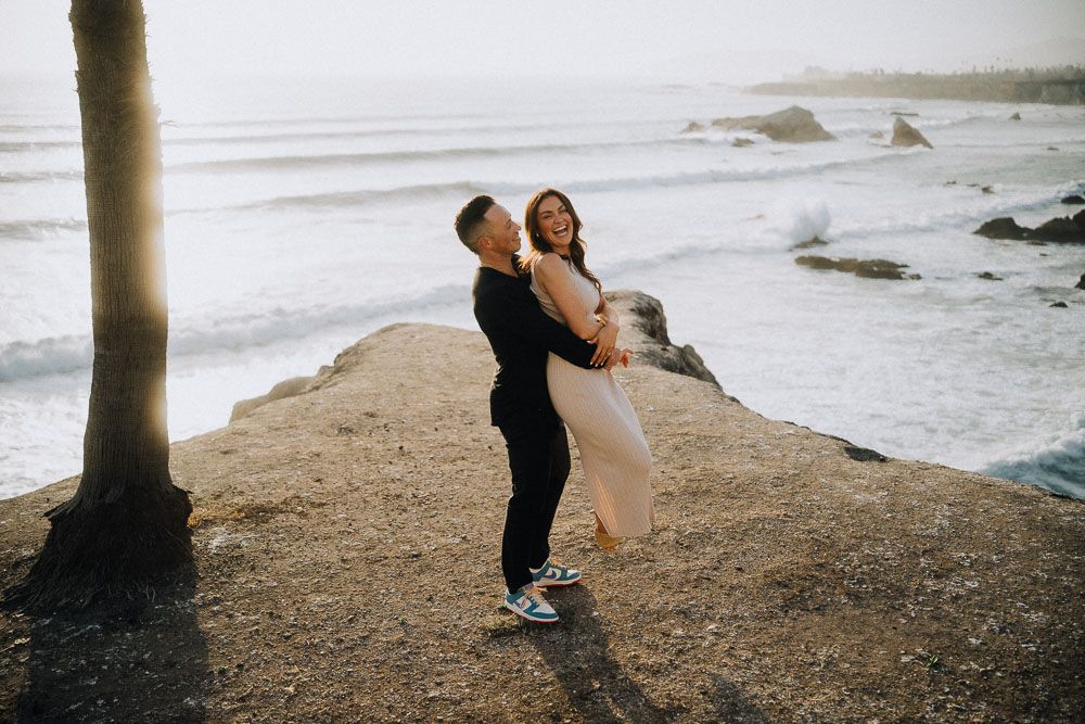 A romantic beach photoshoot at sunset with silhouettes embracing against the ocean and rocky cliffs.