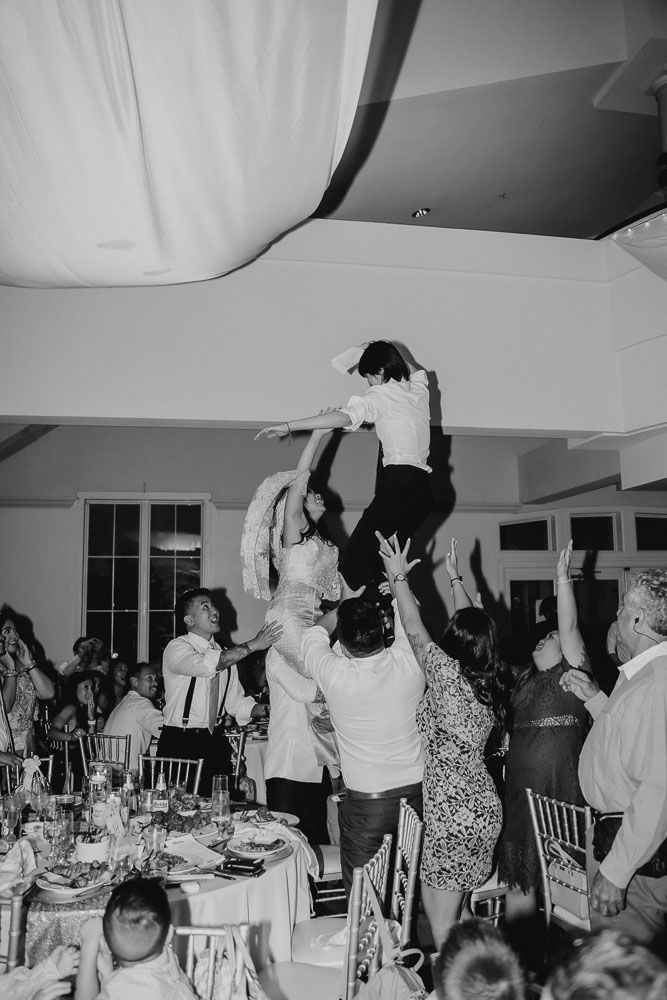 Black and white photo of guests lifting someone high up during an energetic wedding reception moment.