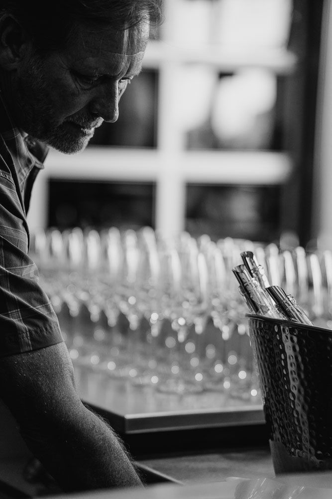Black and white image of a bartender preparing drinks at a well-stocked bar.