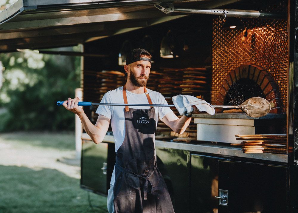 Chef in an apron holding tools near an outdoor pizza oven.