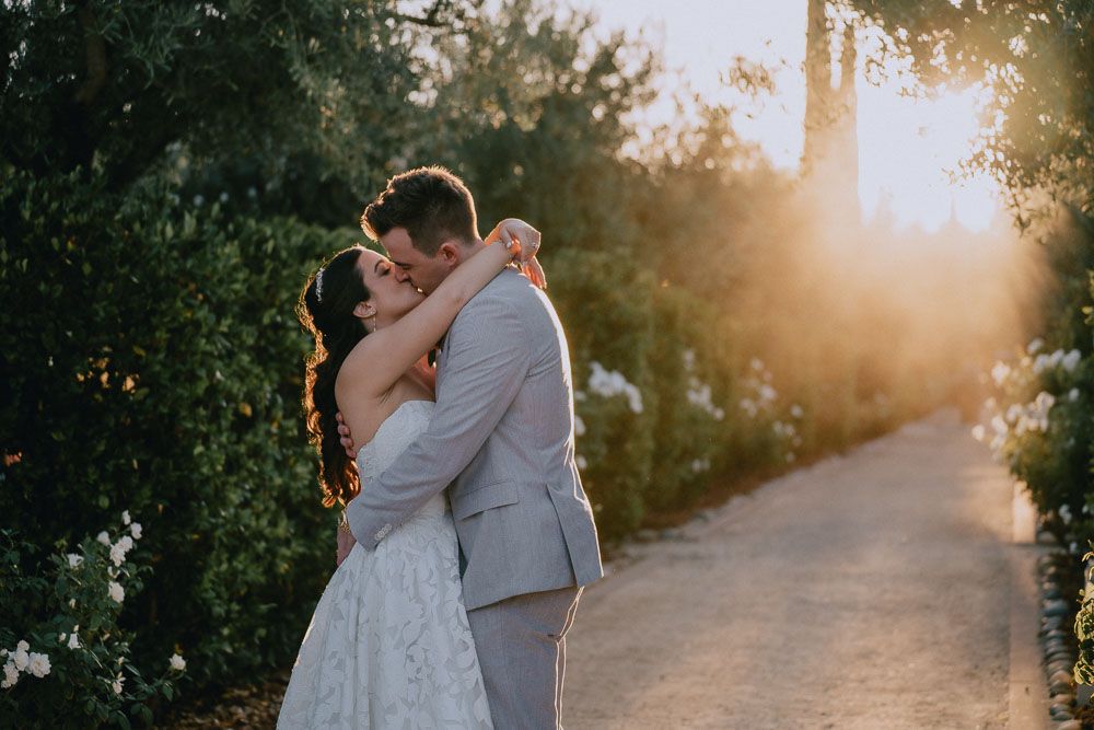 Romantic sunset portrait of newlyweds embracing on tree-lined path.