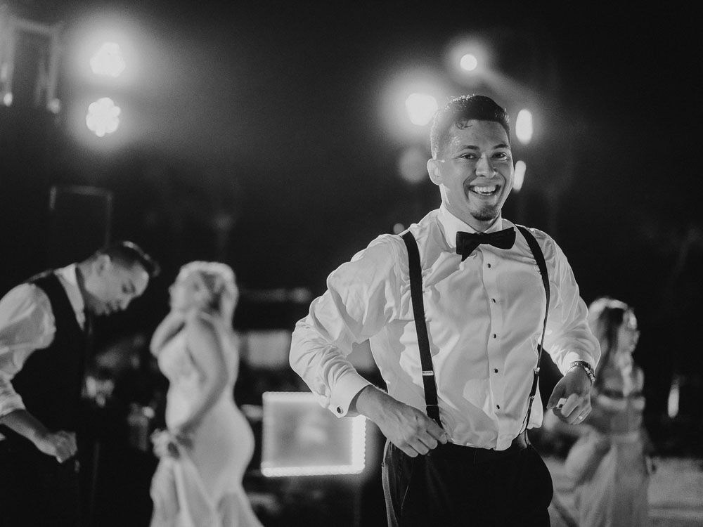 Wedding guest in suspenders and bow tie smiling during evening reception celebrations.