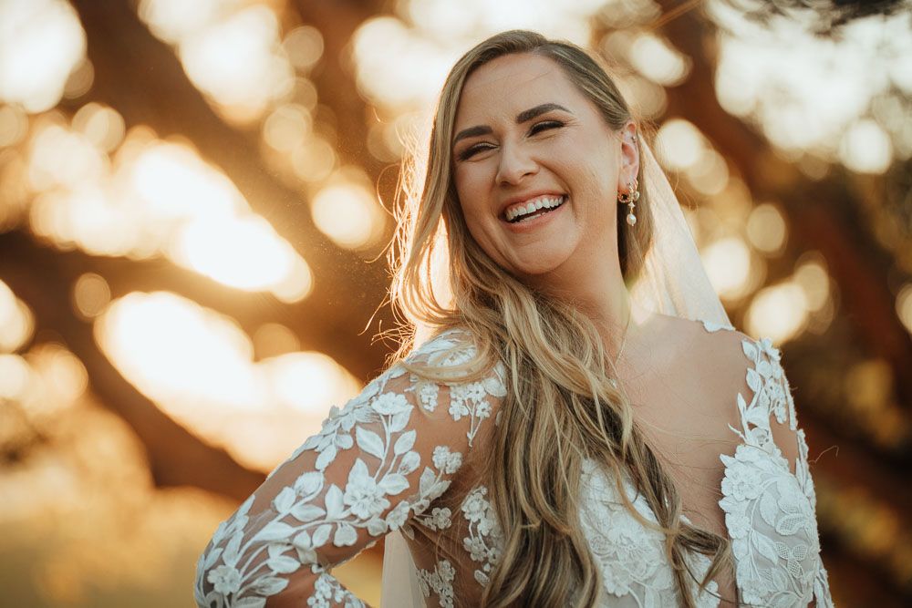 Backlit portrait of bride in lace dress laughing during golden hour.