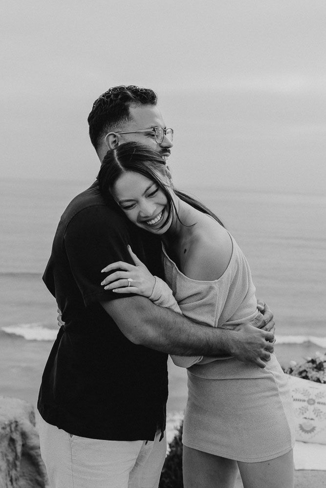 Black and white portrait of embracing couple against ocean backdrop.