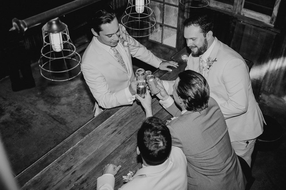 Black and white photo of wedding guests gathered around a wooden table sharing drinks.