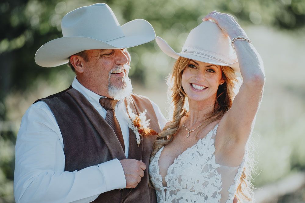Western-themed wedding portrait with couple wearing cowboy hats in sunlit outdoor setting.