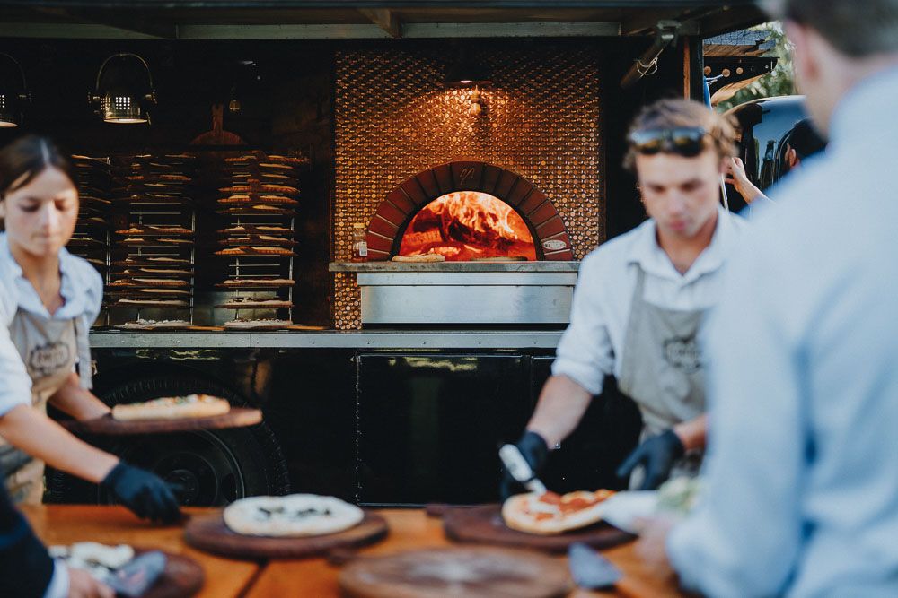 Staff preparing food near a brick wood-fired pizza oven in a restaurant setting.
