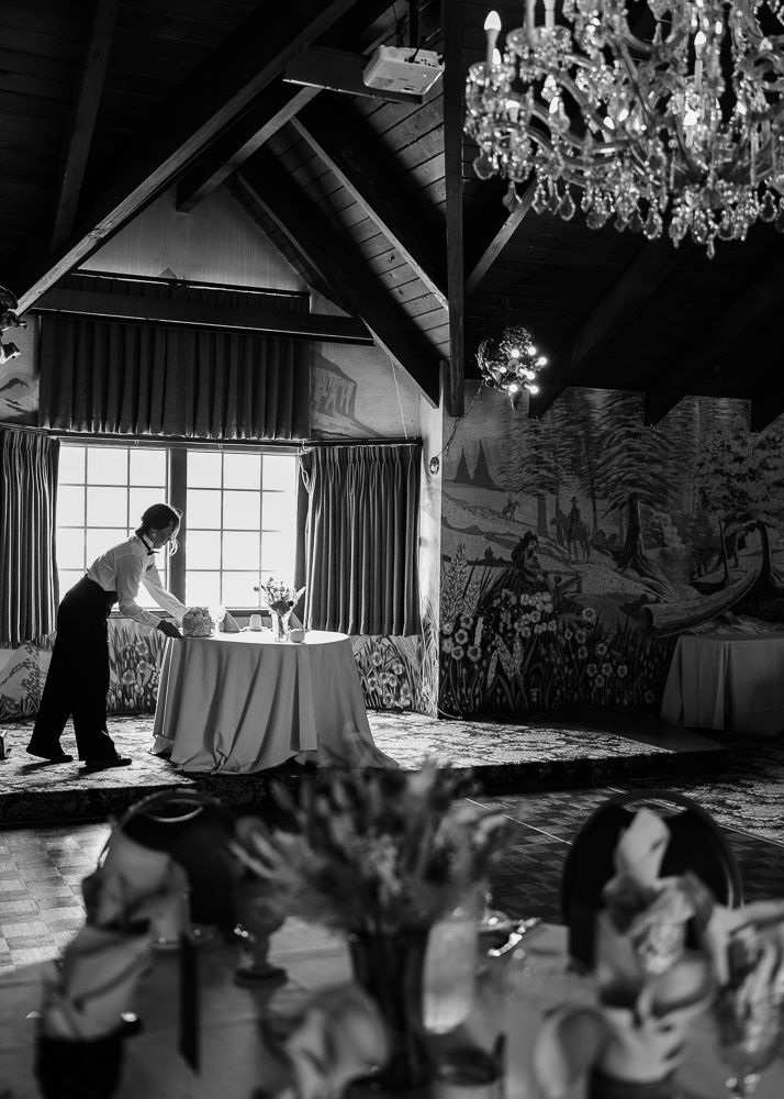 Dining room with chandelier and server setting an elegant table near large windows.