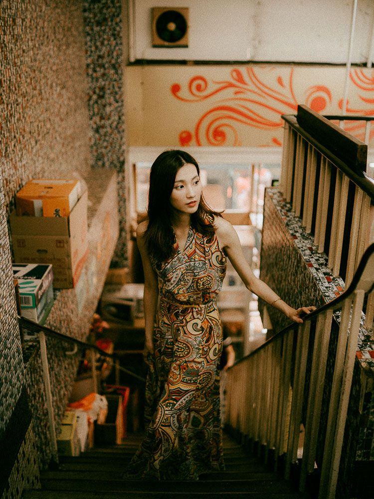 A woman in a patterned dress poses on vintage wooden stairs with decorative orange wall designs.