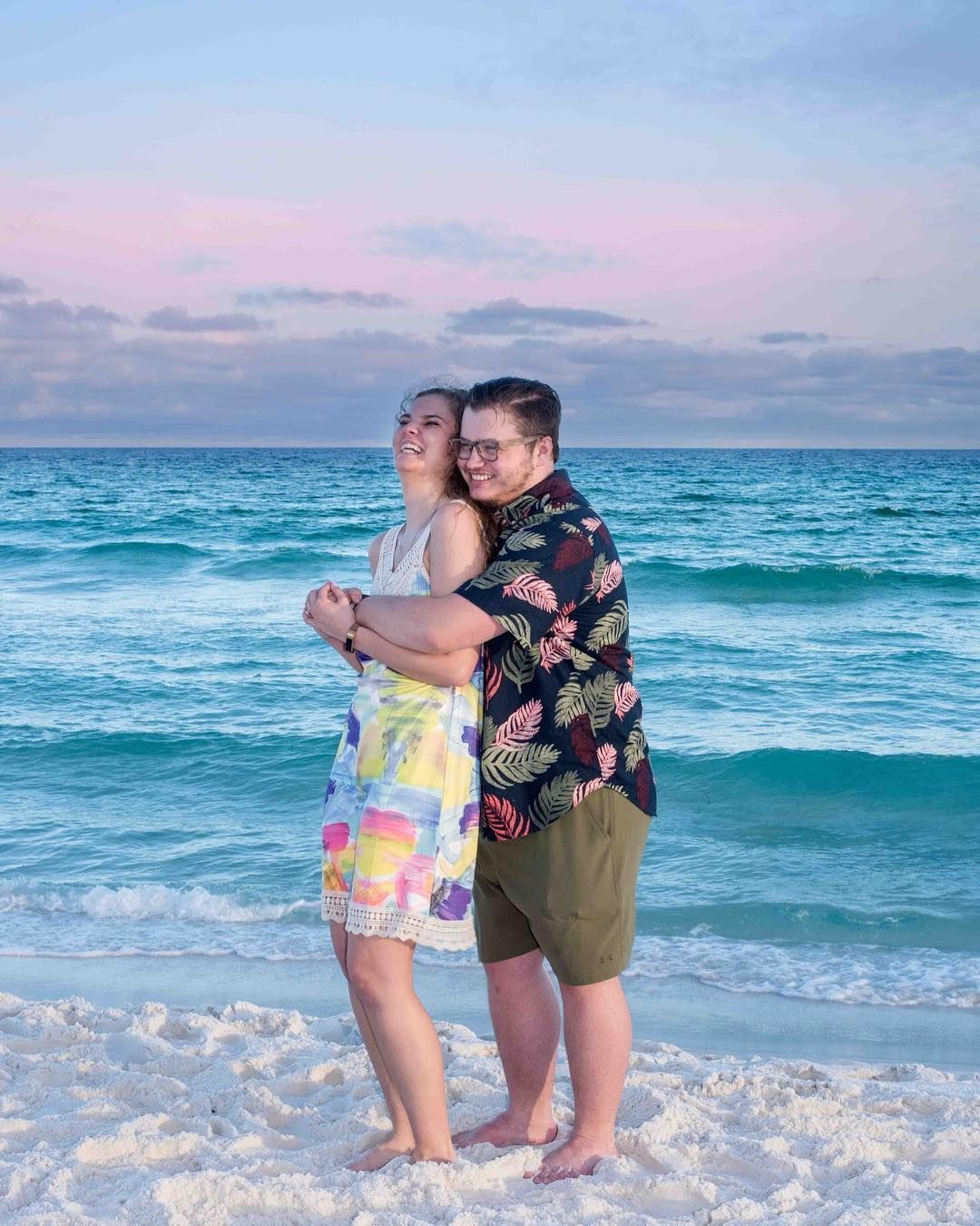 Two people embrace while standing on a beach with turquoise waves and pink sunset sky in the background.