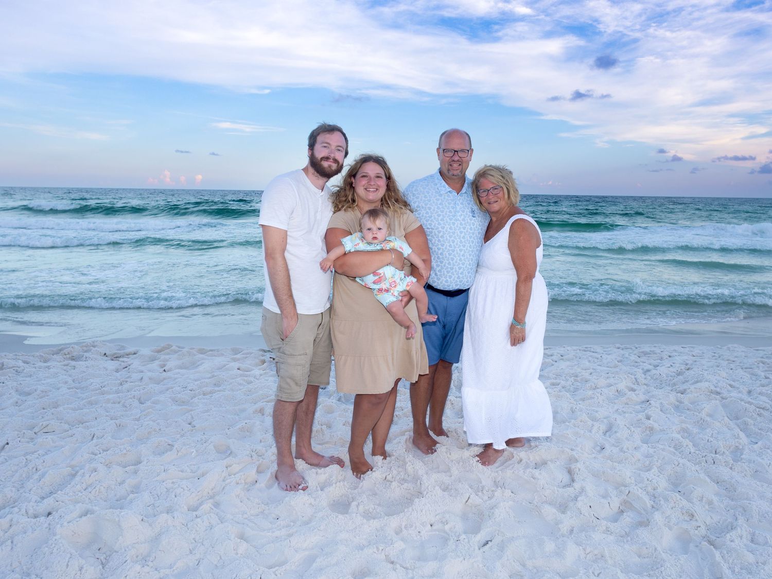 A family group wearing white and khaki clothing poses together on a pristine white sand beach with turquoise waves behind them.