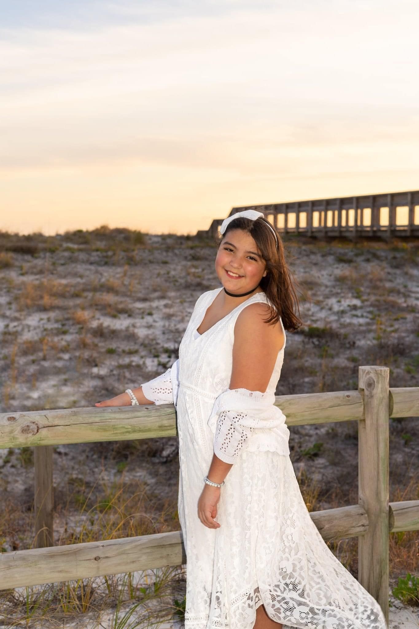 A white lace off-shoulder dress photographed in a coastal setting with wooden railings at sunset.