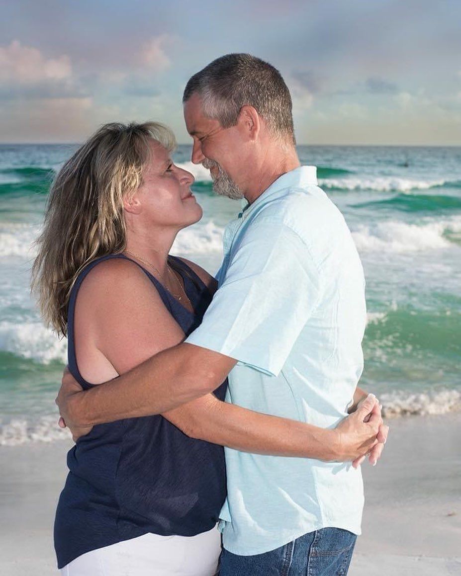 A couple in navy blue and white embrace on a beach with ocean waves in the background during sunset.