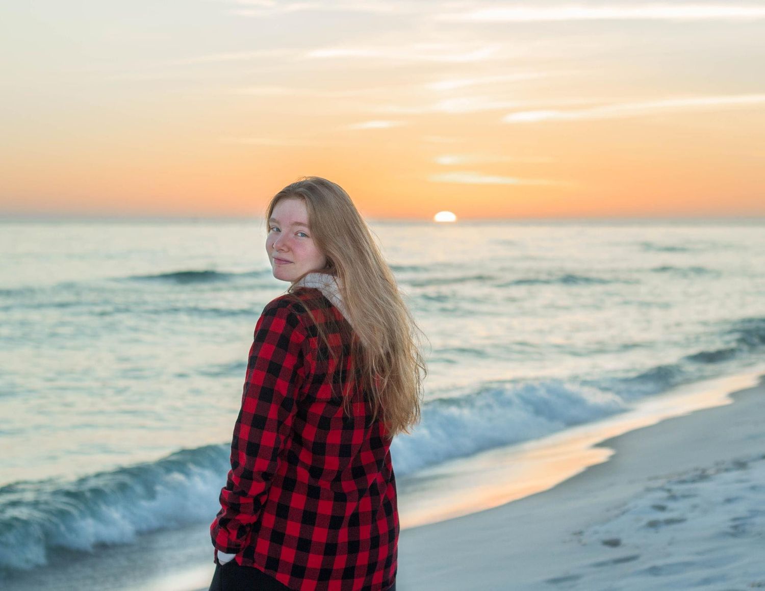 A woman in a red buffalo plaid shirt stands on a beach at sunset as waves crash on the shore.