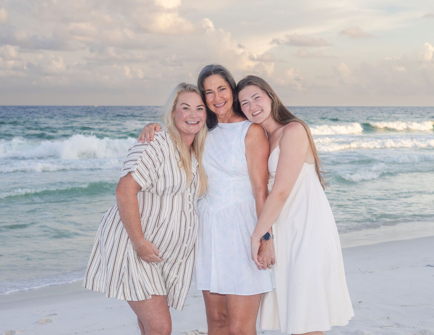 Friends in white outfits embracing on beach with ocean waves in background.