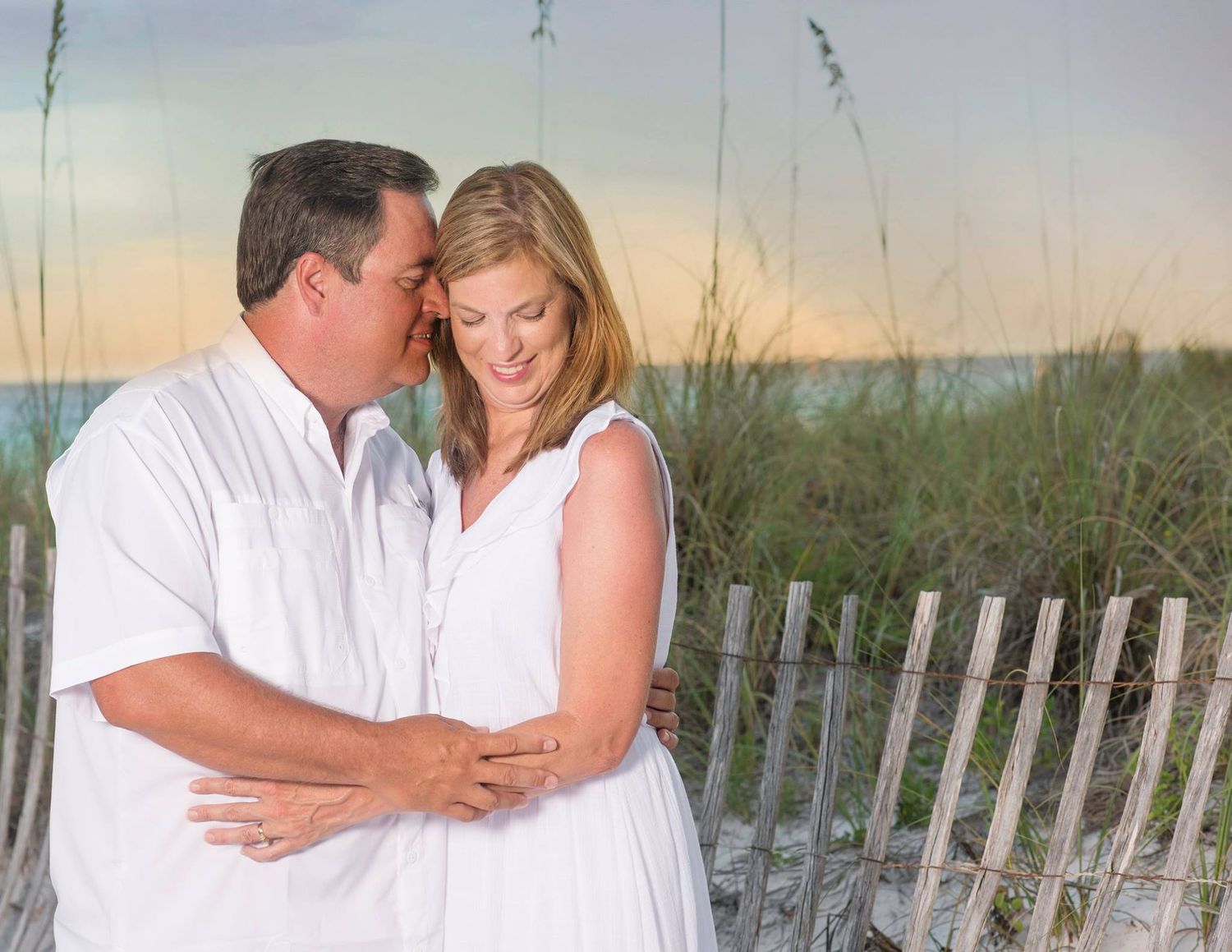 Romantic couple embraces on beach boardwalk with sea grass and sunset sky in background.