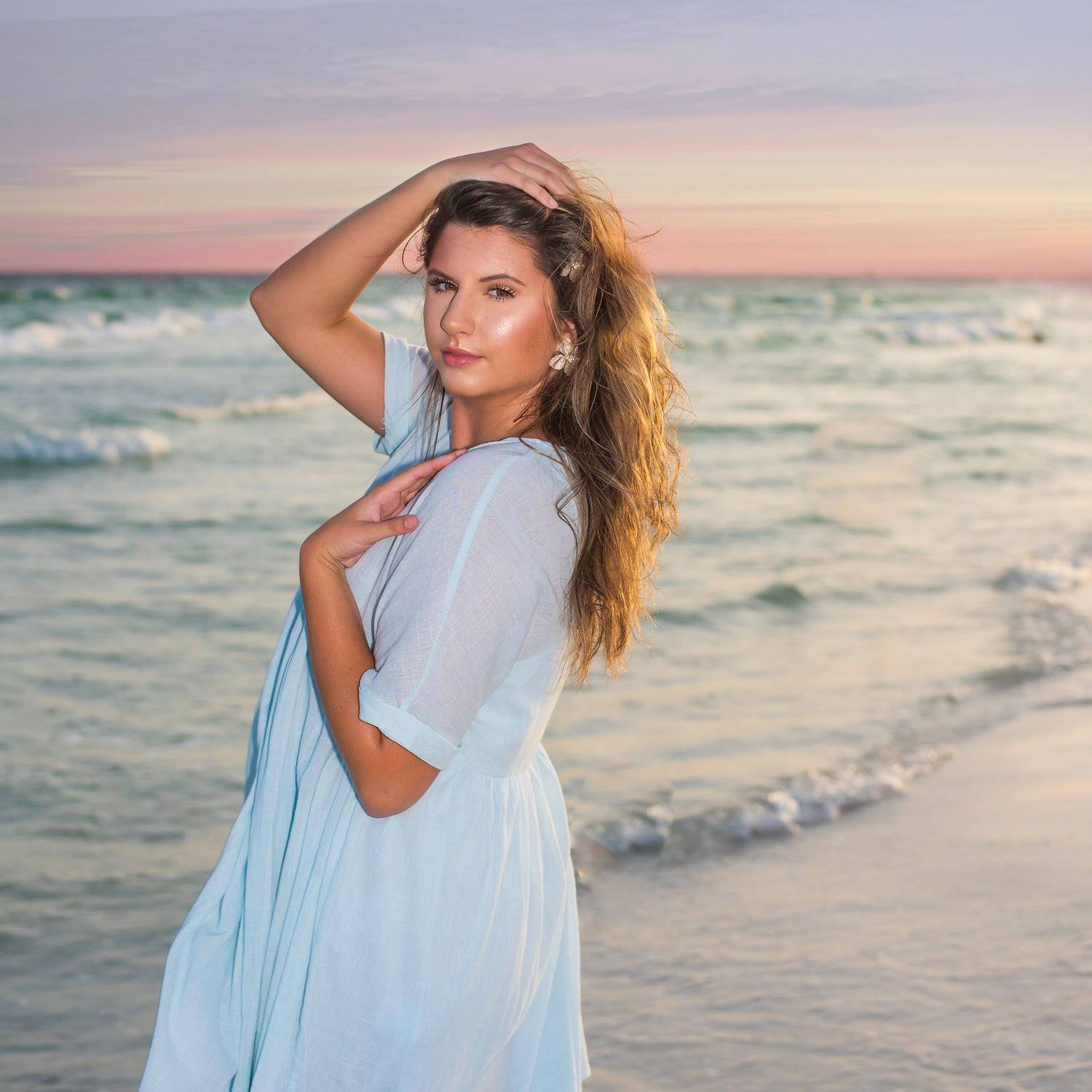 A woman in a white dress poses on a beach at sunset with waves crashing in the background.