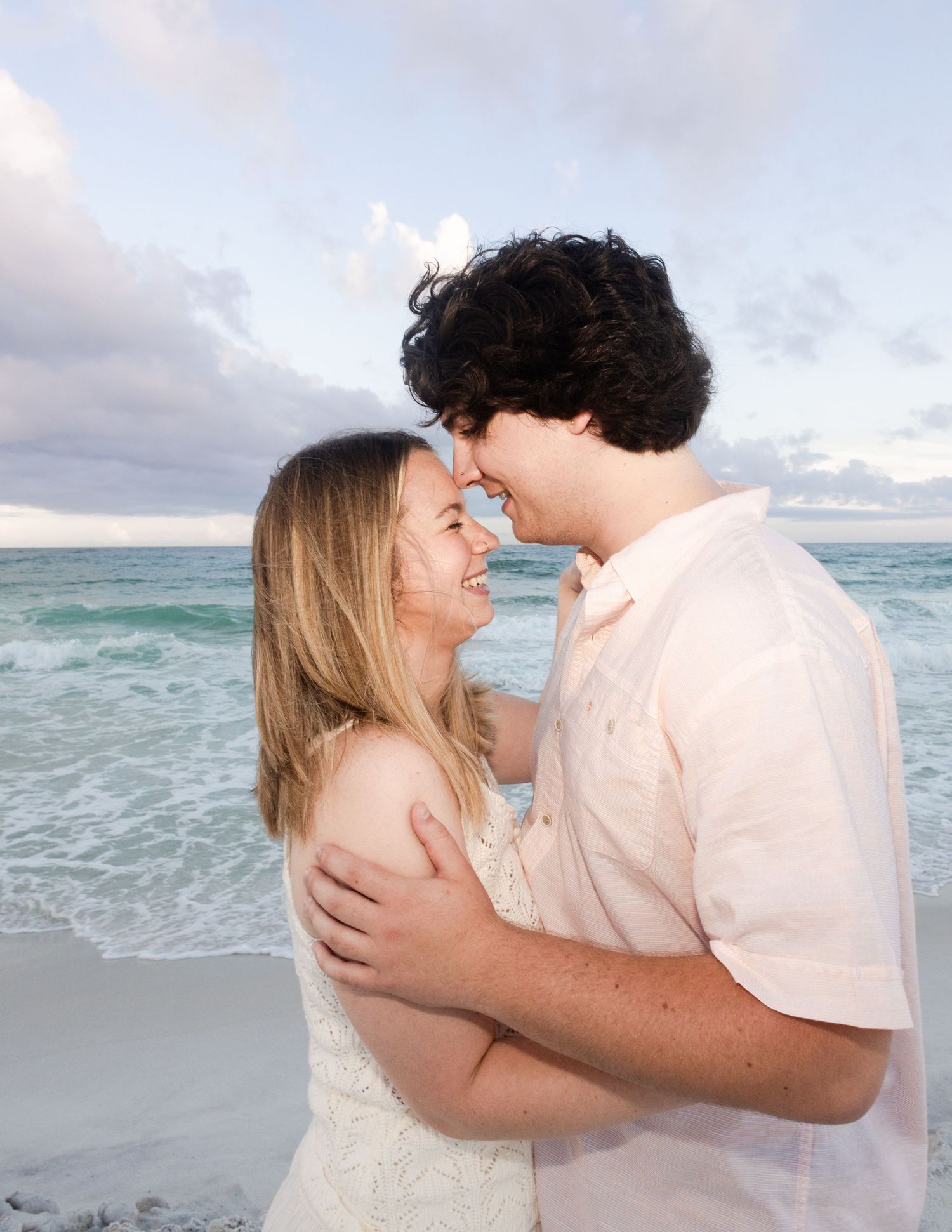 A couple shares a romantic moment on a beach at sunset, smiling and embracing with ocean waves in the background.