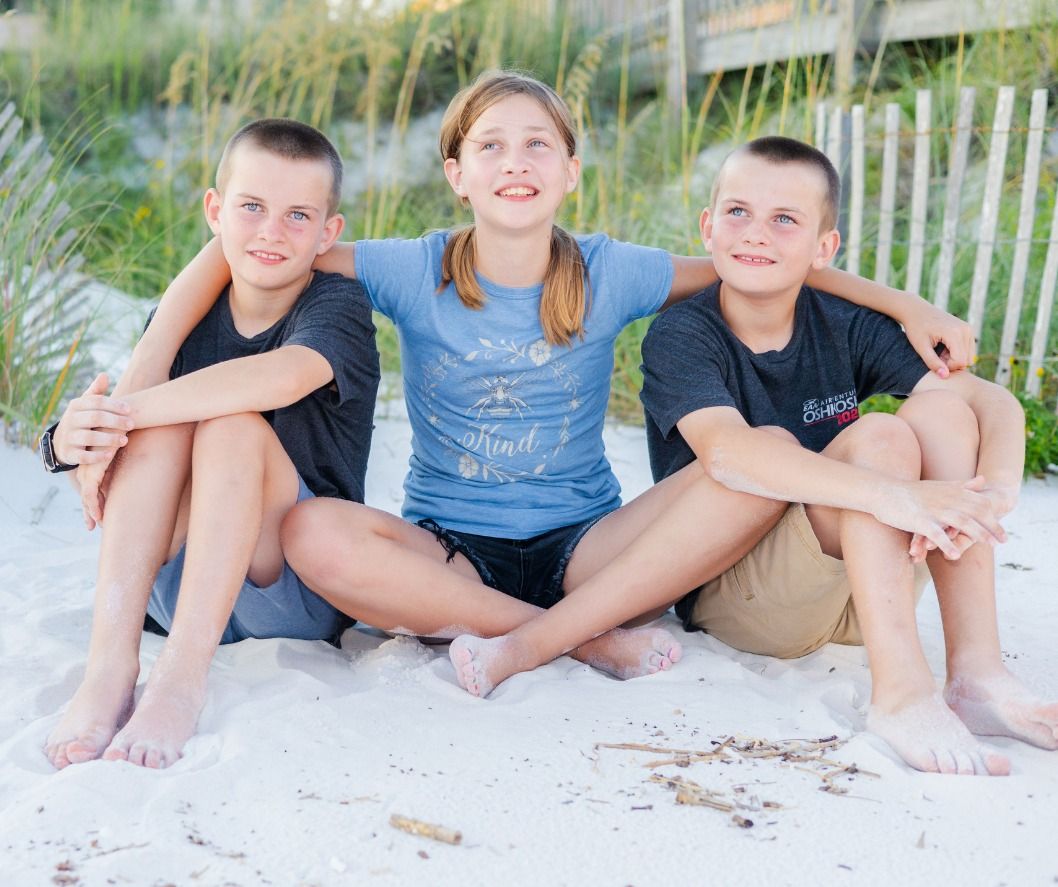 Three children sit together smiling on a sandy beach with sea grass and dunes in the background.