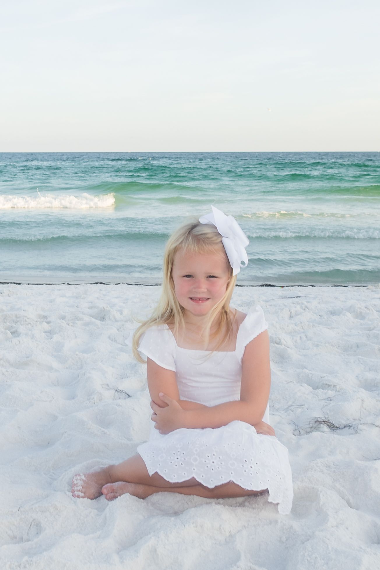 Young girl in white dress lying on pristine beach sand during sunset photoshoot.