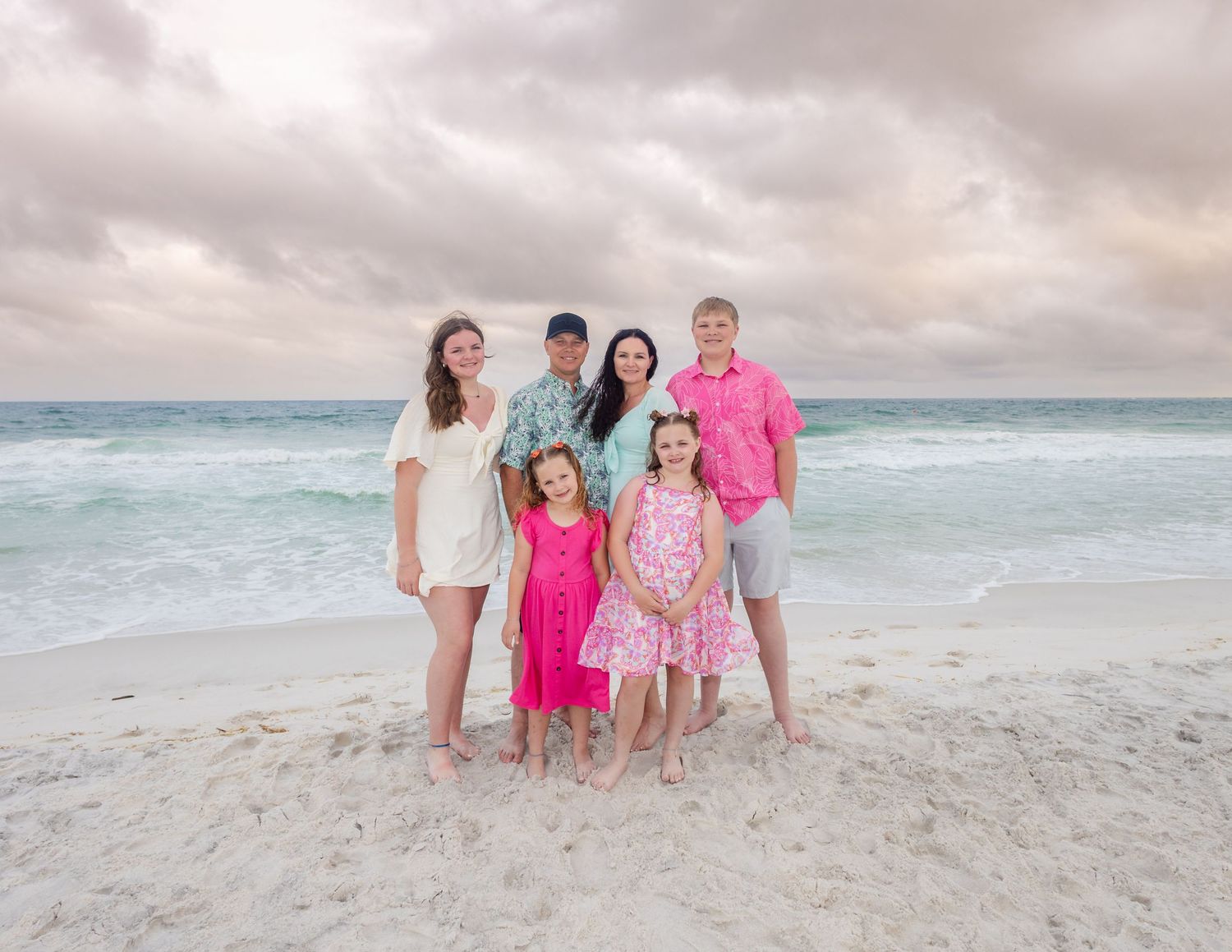 Family of six posing on a sandy beach with turquoise ocean waves and cloudy sunset sky in the background.